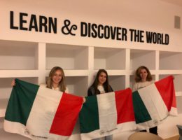 three women each hold an Italian flag underneath a sign that says "Learn and Discover the World"